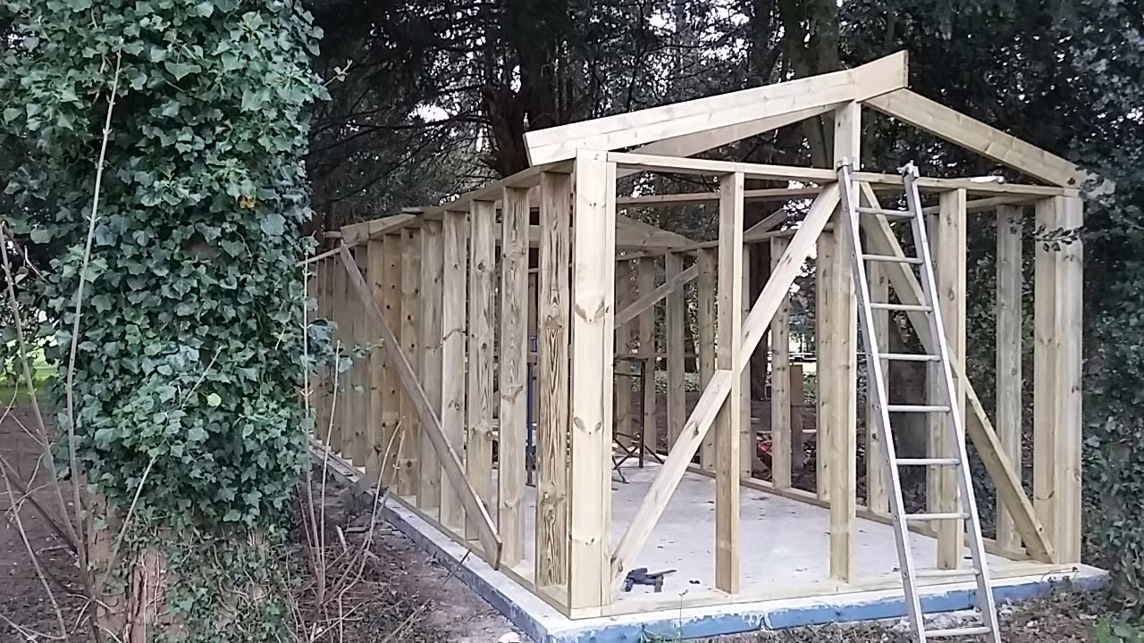 The treated timber frame of a garden building going up on a concrete base, a ladder against the ridge