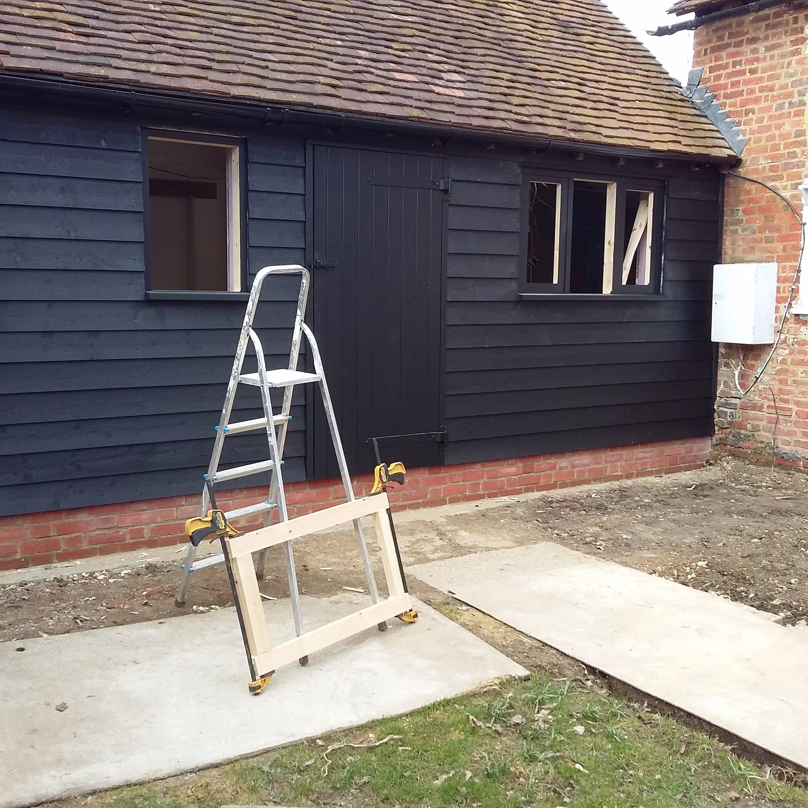 A finished outbuilding in black weatherboard on a red brick plinth, with a ledged door and new window frames